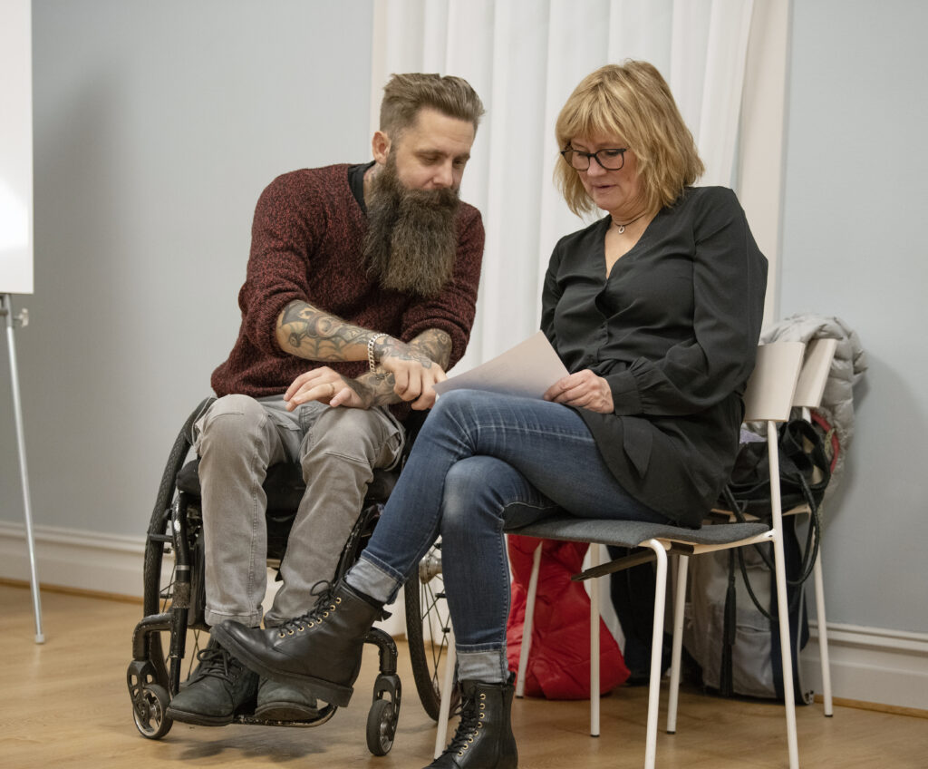 En person i rullstol och en person på vanlig stol pratar om något som står på ett papper