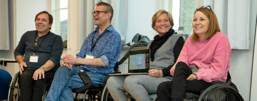 Fyra glada personer varav en har en uppkopplad dator i knät och de tre andra använder rullstol